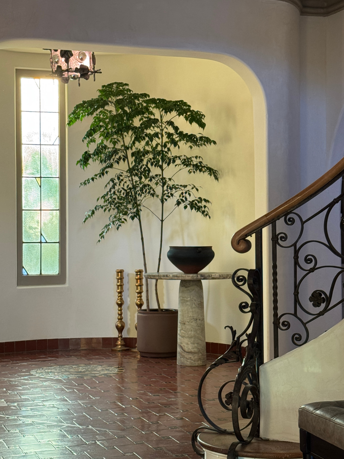 Curved entry with a slender tree, brass candlesticks, and a dark bowl on a travertine table